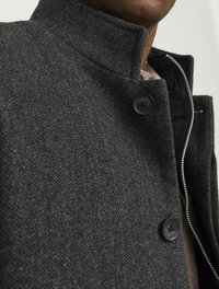 Manteau en chevron gris foncé avec un col haut, doté de boutons noirs et d'une fermeture éclair. Tissu texturé avec une coupe ajustée.
