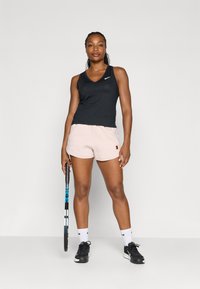 Top deportivo sin mangas negro, pantalones cortos rosas, zapatillas deportivas negras, sosteniendo una raqueta de tenis con acentos azules, de pie sobre un fondo neutro.