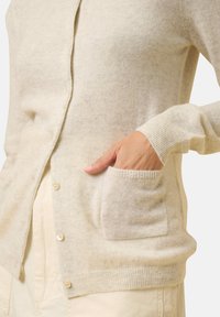 Cardigan beige chiaro realizzato in morbido tessuto a maglia, dotato di scollatura a V, chiusura con bottoni e tasca frontale con polsini a costine.