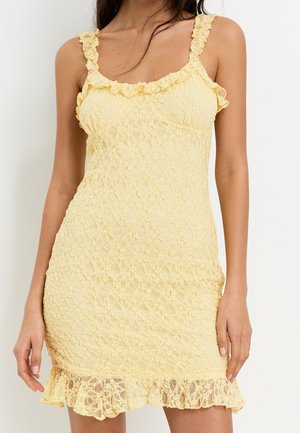 Robe en dentelle jaune avec des bretelles volantées et un motif floral texturé. L'ourlet est terminé par un détail en dentelle froncée. Silhouette ajustée.