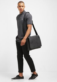 Borsa a spalla grigia scura realizzata in materiale testurizzato, caratterizzata da una chiusura a patta e una tracolla regolabile, portata da una persona in abbigliamento casual.