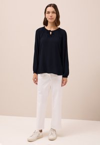 Top blu navy a maniche lunghe con scollatura arricciata e dettaglio a occhiello, abbinato a pantaloni bianchi a gamba larga e sneakers di colore chiaro.