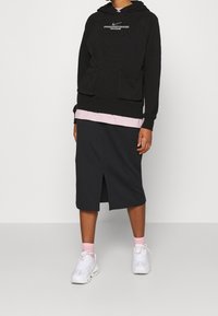 Svart hoodie med framfickor och vit text, lager-på-lager över en ljusrosa topp, ihop med en mörkgrå knälång kjol och vita sneakers.