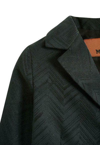 Ciemnozielona marynarka z fakturowanym wzorem w warkocz, z kołnierzykiem typu notch i brązową skórzaną metką przy dekolcie.