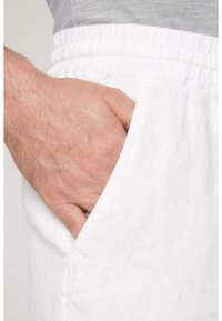 Pantalon en lin blanc avec une ceinture élastique, doté d'une poche latérale. Vue en gros plan mettant en valeur la texture du tissu et une main s'insérant dans la poche.
