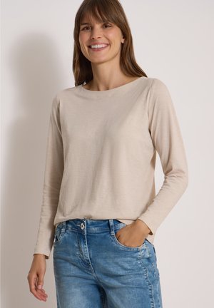 Femme souriante aux cheveux bruns portant une chemise beige à manches longues et un jean bleu, la main droite dans la poche, sur un fond uni.