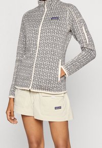 Osoba v šedé bundě na zip s potiskem a béžových šortkách, oboje s malými logy značky Patagonia, na jednobarevném pozadí.