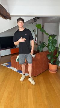 Jauns vīrietis melnā t-kreklā un pelēkos šortos stāv viesistabā ar koka grīdu, rūsganas krāsas dīvānu, lielu zaļu augu un televizoru fonā.