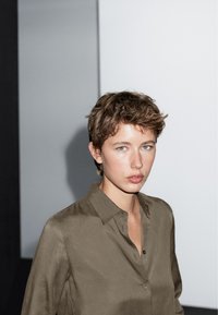 Camicia verde oliva, con bottoni e colletto, dal tessuto morbido. Vestibilità ampia, modello con capelli corti su uno sfondo minimalista.