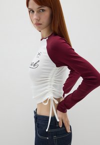 Witte en bordeaux lange mouwen top met zijrouchering, sportlogo opdruk en koorddetail in de taille, gecombineerd met blauwe denim jeans.