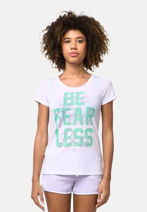 Femme aux cheveux bouclés portant un t-shirt blanc avec l'inscription « BE FEARLESS » en vert et un short violet clair, debout devant un fond uni.