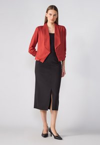 Red, long-sleeved blazer with an open front, paired with a black, fitted dress featuring a front slit. Black high-heeled shoes complete the look.