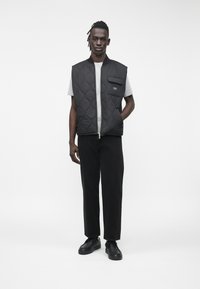 Μαύρο καπιτονέ αμάνικο γιλέκο με φερμουάρ μπροστά και τσέπη στο στήθος, συνδυασμένο με γκρι μπλούζα και μαύρο παντελόνι. Το μοντέλο στέκεται έχοντας τα χέρια στις τσέπες.