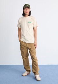 Joven de pie en un suelo azul frente a una pared blanca, vistiendo una camiseta beige de OBEY, pantalones de pana marrones, zapatillas blancas y una gorra de béisbol verde.