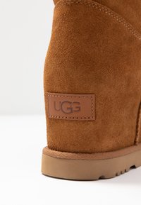 Primer plano del tacón de una bota UGG de ante marrón con parche de logo cosido y suela de goma sobre un fondo blanco.