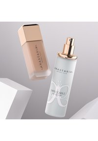 Twee cosmetische producten: een hexagonale foundationfles in beige met een gouden dop, en een doorzichtige setting spray met een gouden spuitkop en een vlinderlogo.