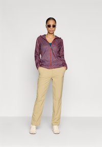 Chaqueta ligera de colores rojo y azul con un estampado, cerrada con cremallera, combinada con pantalones beige y zapatillas beige. Lleva una blusa azul y gafas de sol blancas.