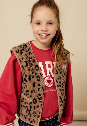 Bruine fleece vest met luipaardprint en een zandkleurige bies, gedragen over een rode sweatshirt met witte tekst. De achtergrond is neutraal beige.