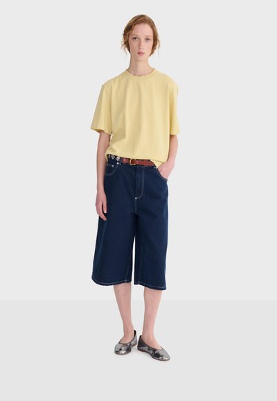 Jeune personne portant une chemise jaune à manches courtes, un pantalon large bleu foncé mi-genou avec une ceinture marron, et des chaussures plates métalliques, debout immobile.