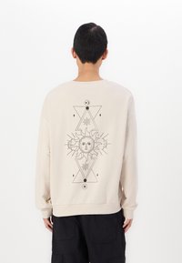 Felpa beige con maniche lunghe, caratterizzata da un design grafico nero di un sole e motivi geometrici sulla schiena. Texture morbida, vestibilità rilassata.