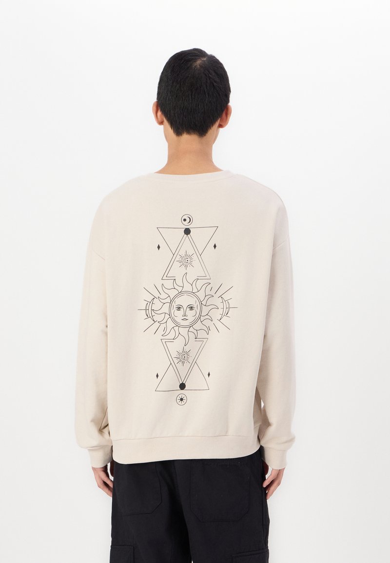 Felpa beige con maniche lunghe, caratterizzata da un design grafico nero di un sole e motivi geometrici sulla schiena. Texture morbida, vestibilità rilassata.