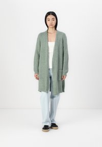Ljusgrönt stickat cardigan med långa ärmar, öppen framsidan och två framfickor, parat med ljusblå jeans och svarta skor.