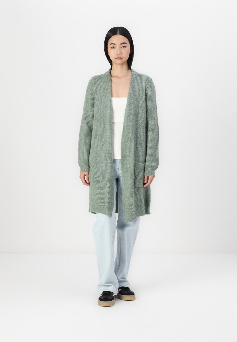 Ljusgrönt stickat cardigan med långa ärmar, öppen framsidan och två framfickor, parat med ljusblå jeans och svarta skor.