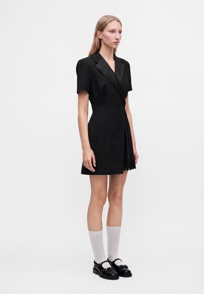 Femme debout portant une robe blazer courte noire à manches courtes, des chaussettes blanches et des mocassins noirs sur un fond blanc uni.