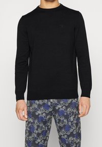 Pull en maille noire avec encolure ronde et manches longues. Présente un détail de logo subtil sur la poitrine ainsi que des poignets et un ourlet côtelés.