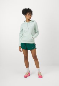 Hoodie verde claro com cordão, bolso frontal e logótipo branco. Combinado com calções de desporto verde escuro e sapatilhas de desporto cor-de-rosa.