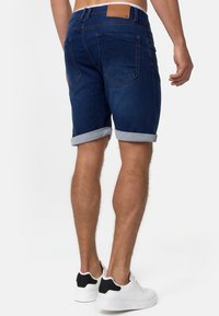 Shorts en denim bleu foncé, avec des ourlets retroussés, des poches arrière et une étiquette en cuir marron sur la ceinture. Portés avec des baskets blanches.