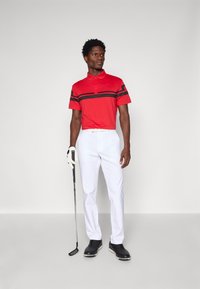 Červené polos tričko s čiernymi pruhmi, biele nohavice a čierne golfové topánky. Drží golfovú palicu a má na sebe bielu golfovú rukavicu.