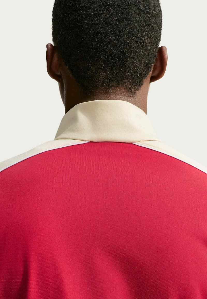 Persona con capelli corti, ricci e neri che indossa una camicia rossa con collo beige, vista da dietro su uno sfondo uniforme.
