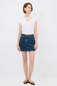 Biała bezrękawnikowa bluzka z detalem w kształcie wycięcia, połączona z granatową dżinsową mini spódnicą. Dodatki obejmują złote bransoletki i brązowe baleriny.