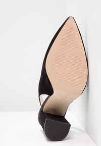 Chaussure à talon haut en daim noir à bout pointu, montrant une semelle beige texturée et un talon bloc, taille 37, placée contre des murs blancs.