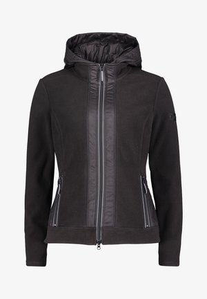 Personne portant une veste à capuche noire zippée avec poches à fermeture éclair, un pantalon noir fuselé, et des chaussures noires faciles à enfiler avec des semelles blanches.