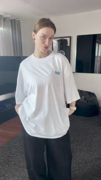 Jovem com cabelo claro preso, vestindo uma camisa branca larga com texto e calças pretas, está de pé numa sala de estar.