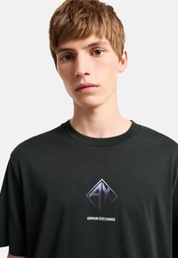 Camiseta de algodón verde oscuro con cuello redondo, con un logo en forma de diamante metálico gris que incluye el texto "AX" y "Armani Exchange" centrado en el pecho.