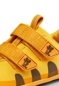 Żółty dziecięcy but z dwoma paskami na rzepy, ozdobiony emblemami z kreskówkowym tygrysem oraz brązowymi wzorami przypominającymi odciski pazurów po bokach.