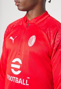 Czerwona koszulka sportowa z pół zamkiem, z teksturowanymi ramionami, logo Puma, okrągłym białym nadrukiem oraz białym emblematem AC Milan.