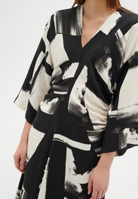 Mustavalkoinen kuvioitu mekko, jossa on V-pääntie, lyhyet hihat ja rypytetty yksityiskohta, valmistettu tekstuurikankaasta, jossa on abstrakteja painatuksia.