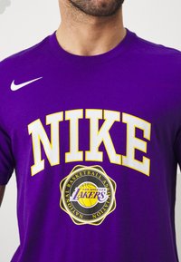 T-shirt Nike violet avec le logo "NIKE" en lettres blanches audacieuses et un logo circulaire des Los Angeles Lakers avec des accents jaunes et noirs.