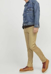 Giacca di jeans con due tasche sul petto, t-shirt marrone, pantaloni slim-fit beige e scarponi da trekking marroni con lacci rossi e occhielli in metallo.