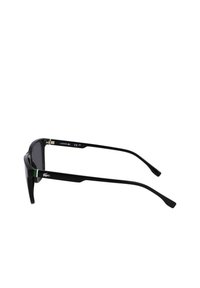 Ochelari de soare negri cu un design elegant, având lentile întunecate și accesorii verzi pe brațe, marcate cu logo-ul Lacoste.