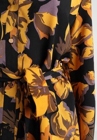 Blommig klänning med stora gula, lila och bruna blommor på en svart bakgrund. Har knappar och ett matchande bälte.