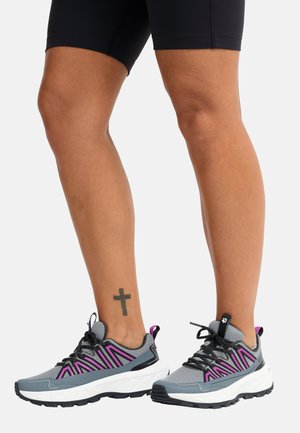 Benen met zwarte shorts en grijze sportschoenen met roze accenten en dikke witte zolen, tegen een effen lichte achtergrond.