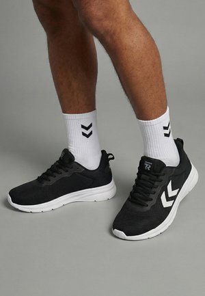 Nogi w białych skarpetkach z czarnymi logo w kształcie jodełki oraz czarne sportowe buty z białymi podeszwami i bocznymi wzorami w kształcie jodełki na szarej podłodze.