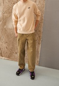 Sweatshirt beige avec un petit logo noir, pantalon cargo olive avec poches latérales, et baskets noires avec des accents violets et le logo.