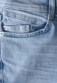 Jeans en denim bleu clair présentant une poche avant avec un bouton de fermeture, des détails de couture visibles et une surface légèrement texturée.