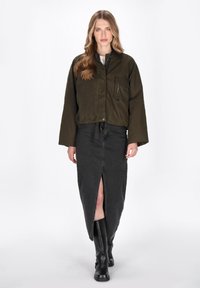 Jachetă scurtă de culoare verde măslinie, cu un buzunar în față și detalii cu fermoar, asortată cu o fustă din denim negru, cu o deschidere pe față și cizme negre până la genunchi.